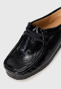 Black patent leather shoe with a rounded toe, elastic laces, and a textured surface. Features a black rubber sole and soft interior lining.