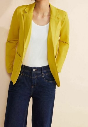 Person iført en knaldgul blazer over en hvid top og mørkeblå jeans, stående mod en ensfarvet lys baggrund.