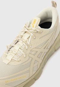 Primo piano di una sneaker ASICS Gel-Quantum 360 beige con dettagli testurizzati, lacci decorati e logo giallo del marchio sulla linguetta e sul lato.