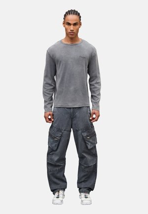 Hombre con cabello trenzado que lleva una camisa de manga larga gris, pantalones cargo oscuros y zapatillas blancas, de pie contra un fondo blanco.