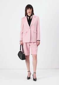 Blazer rosa con due tasche frontali, abbinato a pantaloni corti rosa su misura. Blusa nera sotto, con una borsa nera. Tacchi neri.