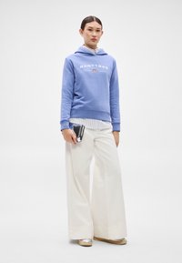 Sudadera con capucha azul con texto blanco "GANT1949", llevada sobre una camisa a rayas, combinada con pantalones blancos de pierna ancha y zapatos plateados. Clutch negro en la mano.