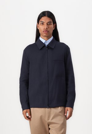 Navy blue zip-up jacket with a collar, featuring a chest pocket, long sleeves, and smooth texture. Paired with beige pants.