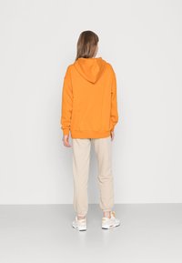 Orange hooded sweatshirt with a loose fit, paired with light beige joggers. Model wearing white sneakers with orange accents.