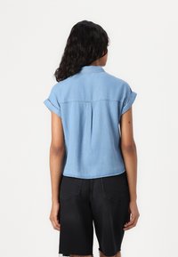 Camisa de ganga azul clara com mangas dobradas, uma pala nas costas e um design encurtado. Combinada com calções pretos com as bainhas desfavoradas.