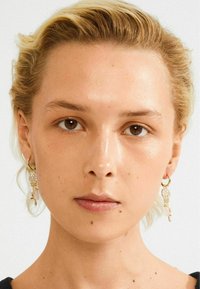 Blonde vrouw met kort haar die gouden oorringen met hangende kristallen draagt, met een directe blik naar voren tegen een effen achtergrond.