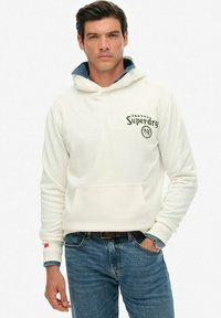 Sudadera blanca con capucha y bolsillo frontal, logo de "Superdry" en verde oscuro, combinada con jeans azules. Presenta un detalle de cuello de mezclilla.
