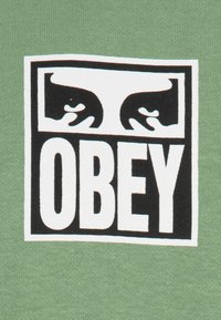 Grön bomullstyg med en vit rektangulär logotyp med svart text "OBEY" och stiliserade svarta ansiktskonturer.