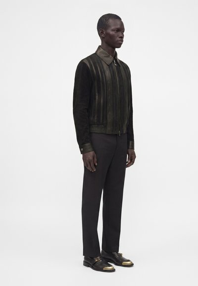 Homme debout portant une veste noire zippée avec des rayures verticales, un pantalon noir et des chaussures noires avec des bouts métalliques dorés.