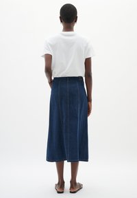 Denim midi krilo v temno modri barvi, z visokim pasom, gladko teksturo in vidnimi šivi. V kombinaciji z enostavno belo majico.
