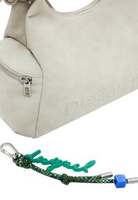 Desigual-Handtasche aus weichem beige Leder mit silbernem Reißverschluss-Detail. Beinhaltet einen grünen Gummischlüsselanhänger mit dem Wort "desigual" und Metallzubehör.
