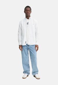 Camicia bianca con bottoni sopra una t-shirt grigia chiara, abbinata a jeans blu larghi e sneakers bianche con dettagli marroni.