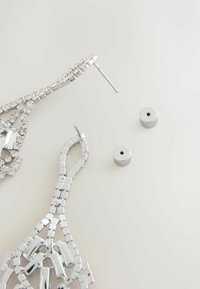 Pendientes de chandelier plateados con diseños intrincados y brillantes rhinestones, acompañados de respaldos de pendientes plateados.