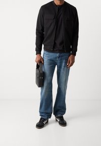 Veste aviateur noire avec deux poches frontales, associée à un jean large bleu et des baskets noires. Tenant un sac rectangulaire noir.