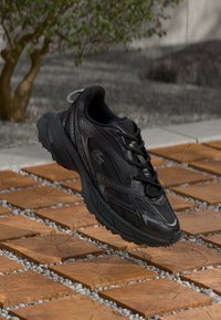 Sorte atletiske sko med teksturerede overflader og buet design, der har snørebånd og gummisåler. Placeret over brune belægningssten.