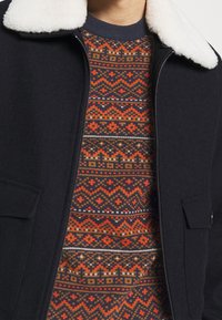 Person som bär en svart jacka med dragkedja och vit fleecekrage över en flerfärgad mönstrad tröja med röda, orangea och bruna geometriska mönster.