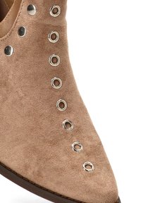 Bota corta de gamuza color canela con punta puntiaguda, que presenta una fila de ojales plateados a lo largo del lateral como acento y detalle.