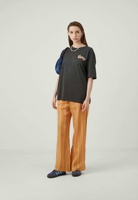 Young woman wearing black graphic t-shirt, orange striped wide-leg pants, navy sneakers, carrying blue shoulder bag, standing against plain background.