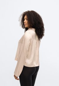 Ανοιχτό μπεζ μπλούζα σε μαλακό, υφασματοποιημένο υλικό, με διακριτό γεωμετρικό σχέδιο και χαλαρή, ελαφρώς κοντή εφαρμογή.