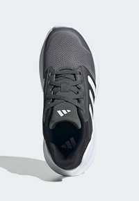 Grauer Sportschuh mit Mesh-Obermaterial, gepolsterter Zunge, schwarzen Akzenten und drei weißen Streifen. Abgerundete Spitze und strukturierte Sohle.
