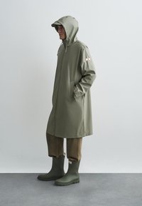 Casaco de chuva longo verde-oliva com capuz, apresentando bolsos laterais e fecho com zíper. Usado sobre calças castanhas e galochas verdes.