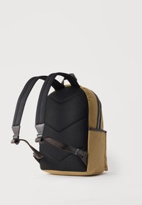 Δίχρωμο καφέ και μαύρο σακίδιο πλάτης με επενδυμένη πλάτη, ρυθμιζόμενους ιμάντες, πλαϊνή τσέπη και φερμουάρ, εμφανιζόμενο από πίσω σε λευκό φόντο.