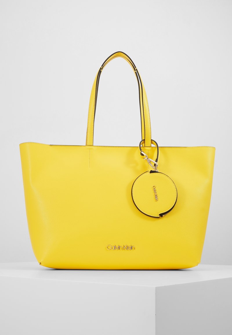 Żółta torba typu tote wykonana z teksturowanego materiału, z dwoma uchwytami, okrągłym breloczkiem oraz logo "Calvin Klein" w metalicznym wykończeniu.