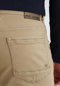 Pantalones de algodón beige con un bolsillo trasero. Presentan un borde cosido y una etiqueta de cuero con la inscripción "PME LEGEND" adherida a la cintura.