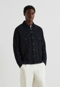 Giacca a scacchi nera realizzata in tessuto testurizzato, con chiusura a bottoni, due tasche sul petto e colletto, abbinata a pantaloni color crema.