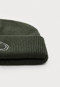 Mørkegrønn strikket lue med ribbestruktur, med en brettet kant og detalj med hvit brodert logo.