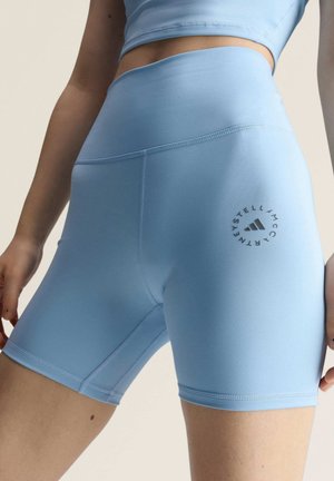 Close-up van een persoon die blauwe, hoog getailleerde sportshorts draagt en een bijpassende crop top met een cirkelvormig logo op de linkerdij.