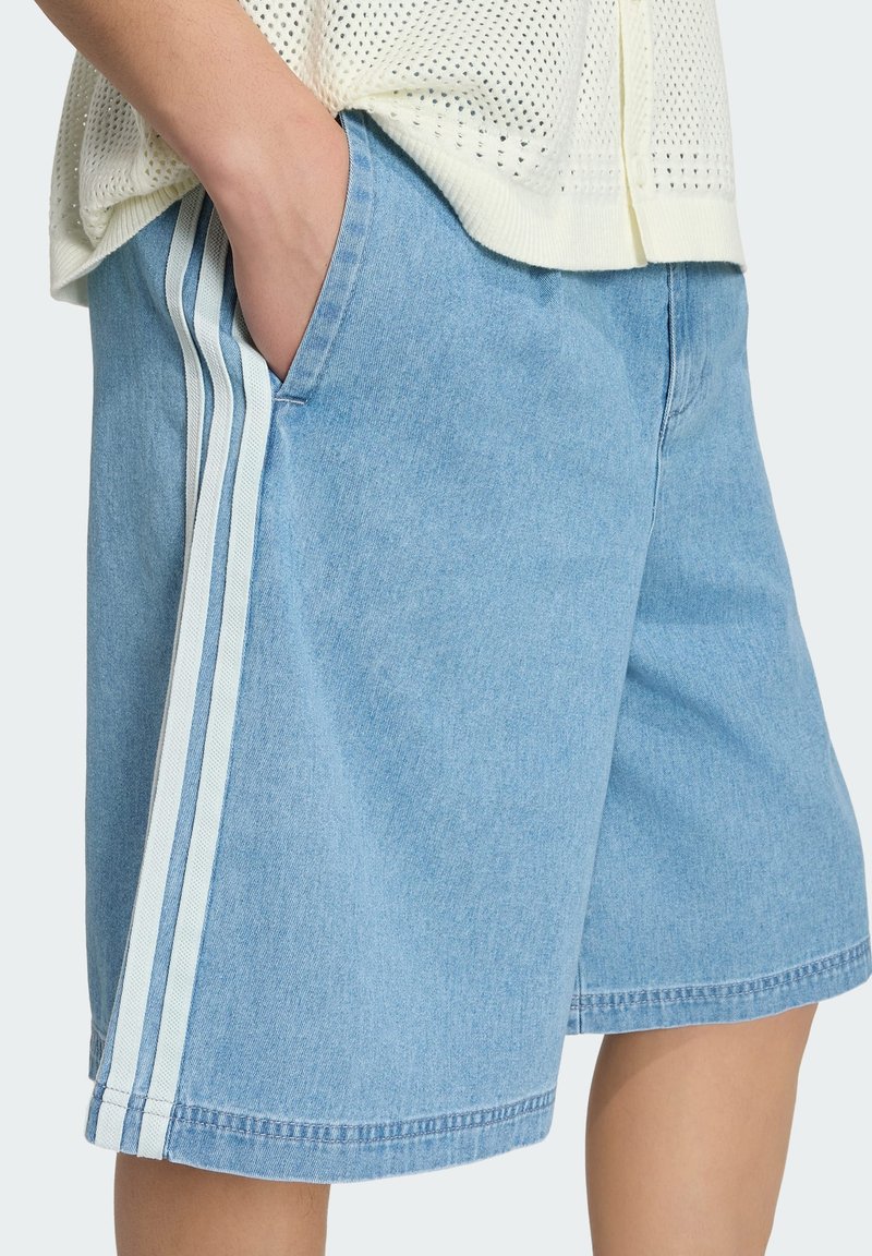 Hellblaue Denim-Shorts mit drei weißen vertikalen Seitenstreifen, Hand in der Tasche, kombiniert mit einem weißen Stricktop.