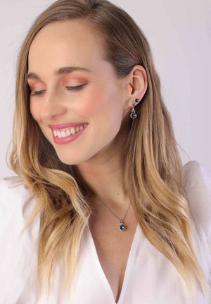 Zilveren sieraden met ronde blauwe edelstenen in oorbellen en een hanger, met een gladde afwerking en een delicate ketting. Haar is golvend.