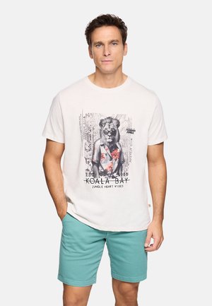 Hombre con camiseta blanca con gráfico de un león con gafas de sol y pantalones cortos verde azulado, de pie con una mano en el bolsillo frente a un fondo liso.