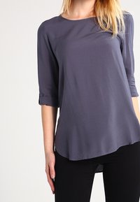 Gray, lightweight blouse with three-quarter sleeves, a round neckline, and rolled cuffs, featuring a slightly curved hem.