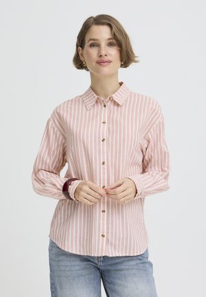 Ženska s kratkimi lasmi, oblečena v roza-belo črtasto srajco z gumbi, svetlo modre kavbojke, zlati uhani in temna zapestnica, stoji pred belim ozadjem.