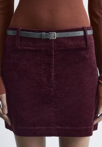 Burgunderfarbene Cordrock mit einer taillierten Passform, ausgestattet mit einem grauen Ledergürtel. Betont durch vertikale Rippstruktur und mit Fronttaschen.