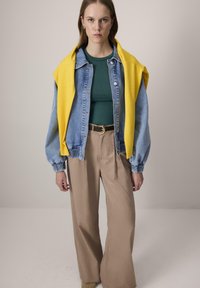 Noor naine kannab rohelist särki, sinist teksajakki, kollast kampsunit õlgadel, beeže laia jalaga pükse, seistes ühtlase tausta ees.