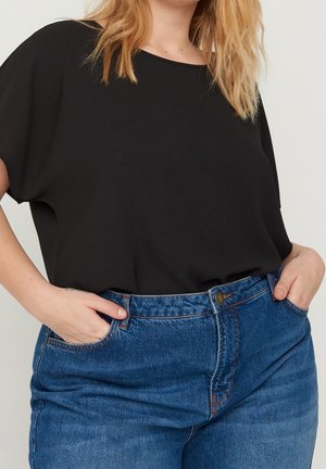 Personne aux cheveux blonds portant un haut noir ample à manches courtes rentré dans un jean bleu taille haute avec les mains dans les poches.