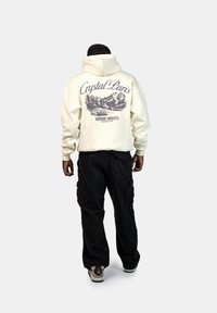 Felpa crema con una grande grafica sul retro che raffigura monti e la scritta "Crystal Paris Nordic Nights." Abbinata a pantaloni cargo neri e sneakers.