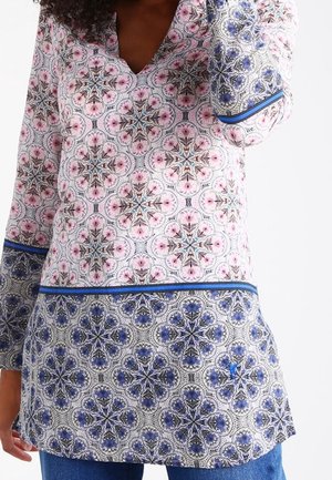 Kvinde iført en langærmet tunika med lyserøde og blå blomster-geometriske mønstre og blå jeans, sat op mod en ensfarvet hvid baggrund.