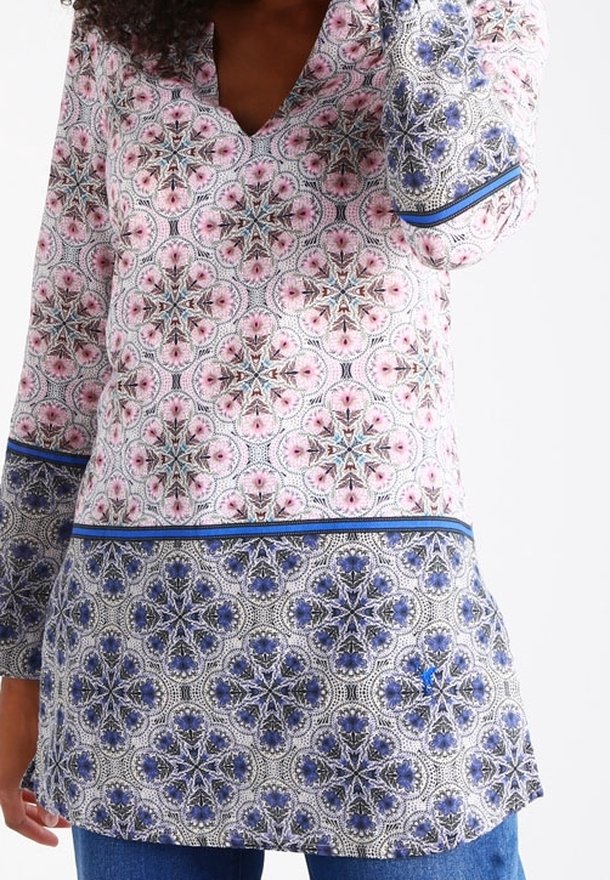 Femme portant une tunique à manches longues avec des motifs géométriques floraux roses et bleus, ainsi qu'un jean bleu, sur un fond blanc uni.