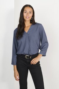 Blusa blu a pois con scollo a V e taglio morbido, abbinata a jeans skinny neri e una cintura con dettagli argentati. Tessuto liscio, maniche lunghe.