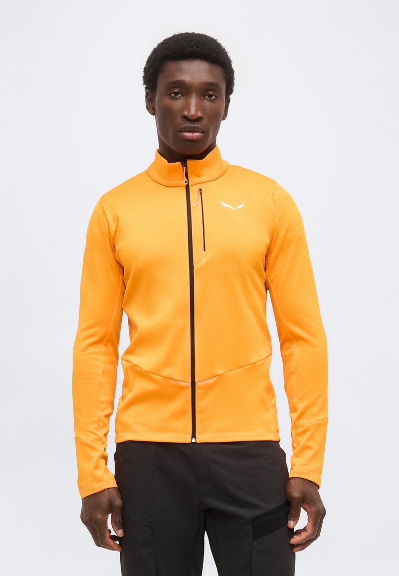 Άνδρας που φοράει φωτεινό πορτοκαλί μπουφάν με φερμουάρ και μικρό λευκό λογότυπο στο στήθος και μαύρο παντελόνι, στέκεται μπροστά σε μονόχρωμο λευκό φόντο.