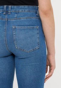 Sinisiä denim-farkkuja, joissa on korkea vyötärö, takatasku ja vaaleaa ommelta. Kangas näyttää sileältä ja siinä on hieman haalistunut pinta.