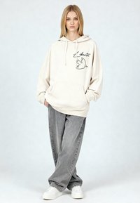 Felpa con cappuccio color crema con tasca a marsupio, presenta il contorno nero di una colomba e la scritta "liberte". Abbinata a jeans grigi larghi e sneakers bianche.