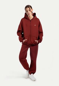 Bordeauxroter Kapuzenpullover mit Reißverschluss, Kapuze und Taschen vorne, kombiniert mit passenden Hosen. Glatte Textur, lockere Passform und weiße Turnschuhe.