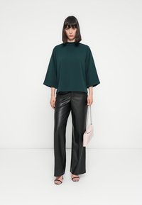 Maglietta oversize verde scuro con maniche larghe, abbinata a pantaloni in pelle nera a vita alta. Borsa rosa e sandali neri con cinturini.