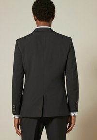 Veste de costume noire cintrée avec une fente à l'arrière, présentant des lignes nettes et un tissu lisse. Les poignets de chemise sont visibles avec des rayures bleues.
