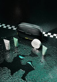 Borsa cosmetica nera con cerniera e accenti gialli, quattro prodotti per la cura della pelle in tubi bianchi e verdi, posizionati su una superficie nera testurizzata con liquido blu.